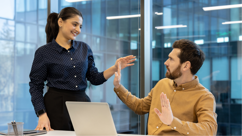 /_cms/webroot/In einem Büro beugt sich eine stehende Person über eine sitzende Person und greift nach ihr, während die sitzende Person mit erhobener Hand ablehnend reagiert.
